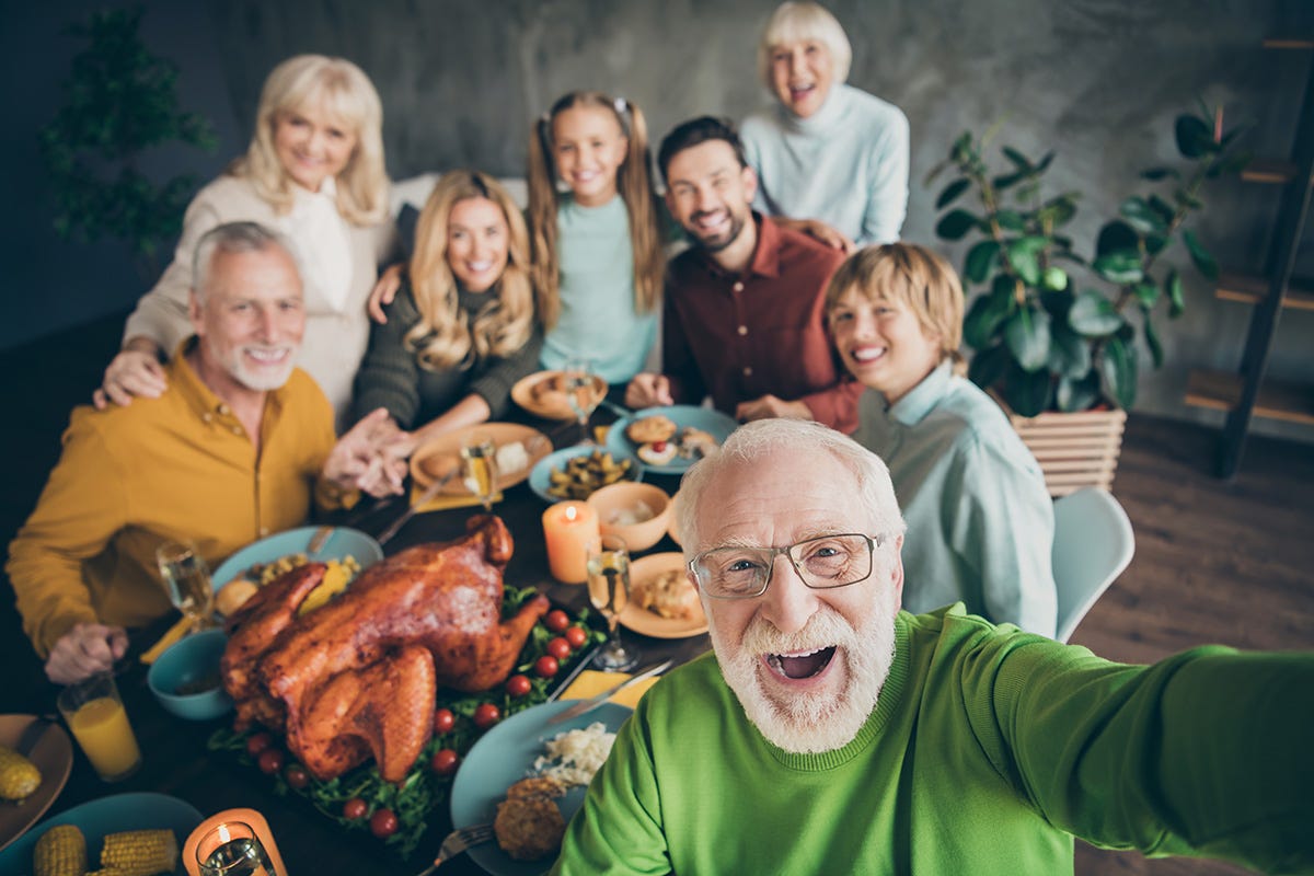 A multi-generational family smiles while posing for a group selfie around the table filled with their holiday feast.