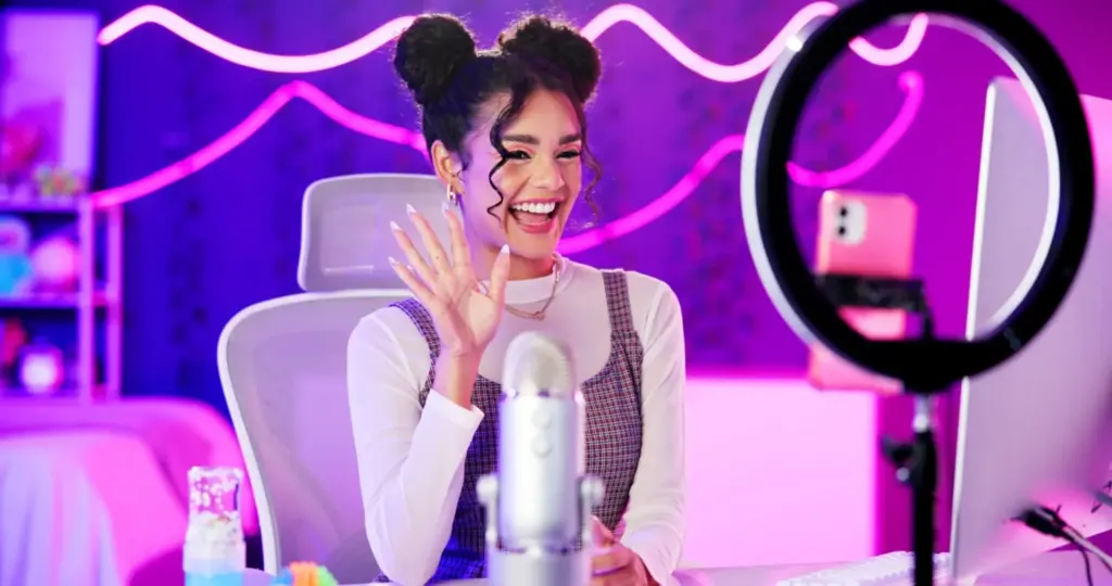 A cheerful content creator with styled hair waves at the camera while recording in a vibrant studio setup with neon lights and a ring light.